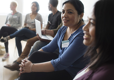 A support group meeting. Participants wear a rectangular name tag on their shirts. A woman with dark hair and eyes and light brown skin is the focus of the photo. She looks off to someone or something not visible in the image, as if she's listening or speaking to them.